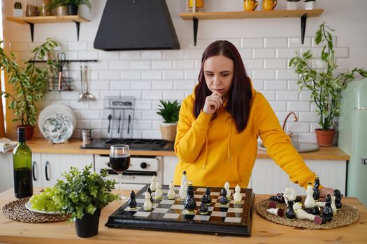Young woman playing chess on kitchen table. Female plays in logical board game with herself in kitchen.