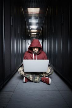 The hacker in a hood sits with the computer among servers, the burglar. Storage of data, cloudy services, system breaking, attack of hackers