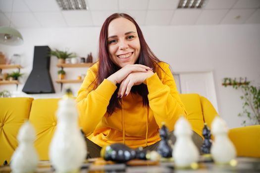 Young woman sitting on yellow sofa and playing chess in room. Female playing in logical board game with herself.
