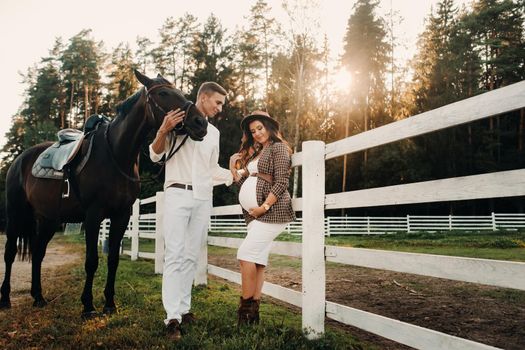 a pregnant girl in a hat and a man in white clothes stand next to horses near a white fence.Stylish pregnant woman with a man with horses.Married couple.