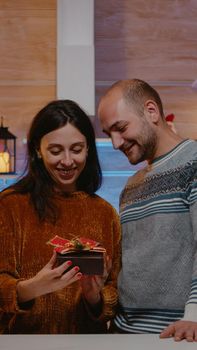 Man giving present to woman for christmas celebration in festive decorated kitchen. Person feeling surprised receiving gift from partner for holiday festivity in winter season. Cheerful couple