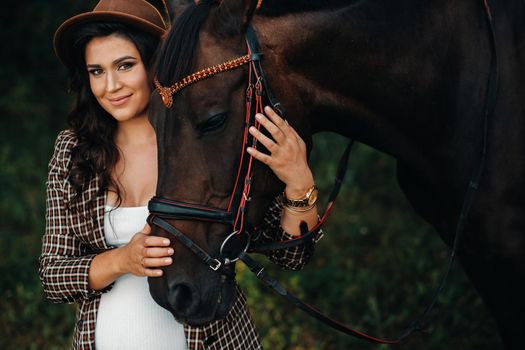 pregnant girl with a big belly in a hat next to horses in the forest in nature.Stylish girl in white clothes and a brown jacket.