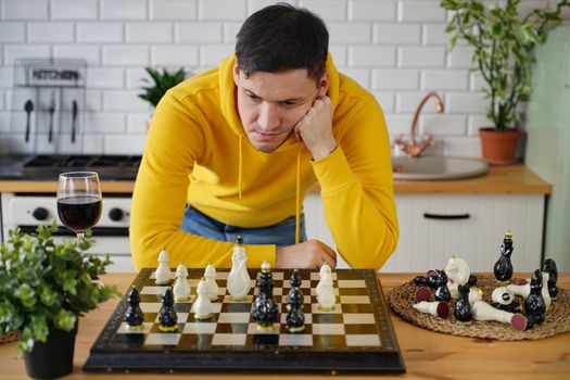 Young man playing chess on kitchen table. Male plays in logical board game with himself, standing in kitchen.