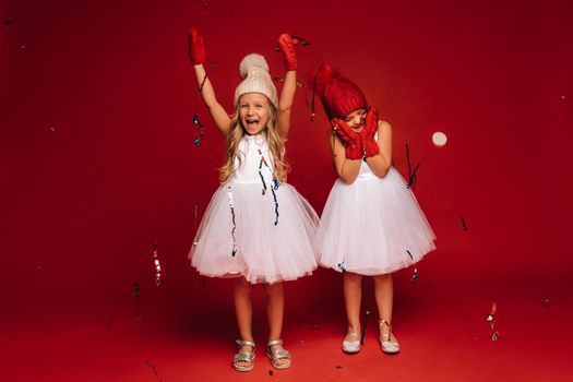 two little girls in white dresses hats and mittens sprinkle confetti on a red background