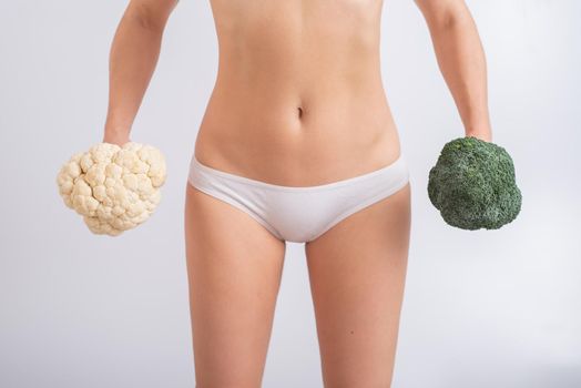 A faceless woman in panties holds cauliflower and broccoli on a white background. Food habits.