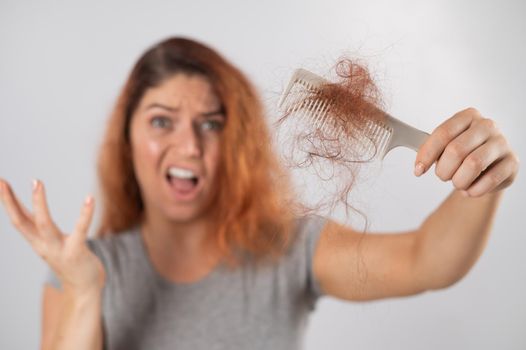 Caucasian woman with a grimace of horror holds a comb with a bun of hair. Hair loss and female alopecia.
