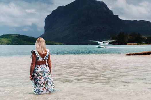 A girl in a swimsuit stands in the ocean and waits for a seaplane against the background of mount Le Morne on the island of Mauritius.A woman in the water looks back at a plane landing on the island of Mauritius