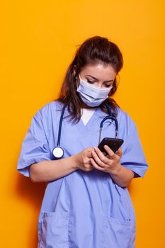 Caucasian nurse looking at smartphone with technology, wearing face mask against coronavirus. Medical assistant with protection and stethoscope working on mobile phone during pandemic