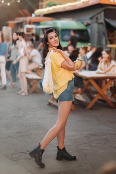 a girl in a yellow shirt, shorts and black shoes with a net of lemons and lemon juice is walking along a city street. A woman with a cocktail in her hands