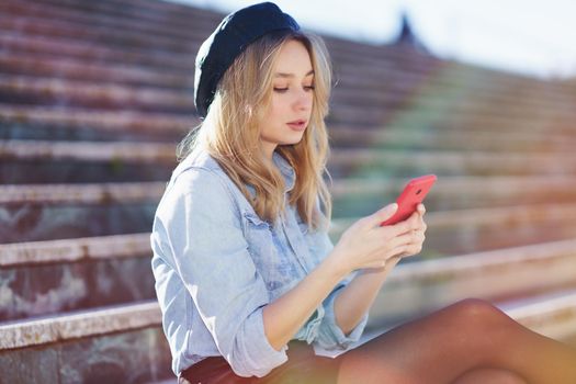 Blonde woman using a smartphone sitting on some city steps, wearing a denim shirt and black beret.