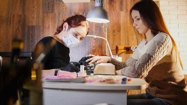 Nail master in medical mask doing professional manicure for white caucasian model, cosmetic salon