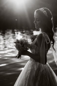 An elegant bride in a white dress and gloves stands by the river in the Park with a bouquet, enjoying nature at sunset.A model in a wedding dress and gloves in a nature Park.Belarus. black and white photo.