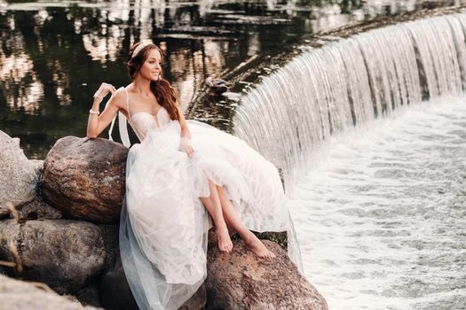 An elegant bride in a white dress, gloves and bare feet is sitting near a waterfall in the Park enjoying nature.A model in a wedding dress and gloves at a nature Park.Belarus