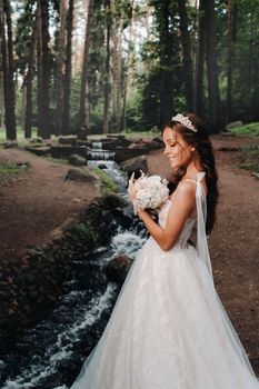 An elegant bride in a white dress and gloves holding a bouquet stands by a stream in the forest, enjoying nature.A model in a wedding dress and gloves in a nature Park.Belarus