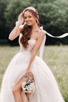 On the wedding day, an Elegant bride in a white long dress and gloves with a bouquet in her hands stands in a clearing enjoying nature. Belarus.
