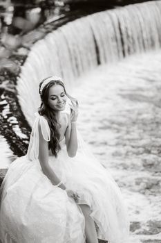 An elegant bride in a white dress, gloves and bare feet is sitting near a waterfall in the Park enjoying nature.A model in a wedding dress and gloves at a nature Park.Belarus
