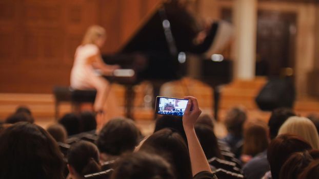 Audience in concert hall during performing piano girl- people shooting performance on smartphone, music opera