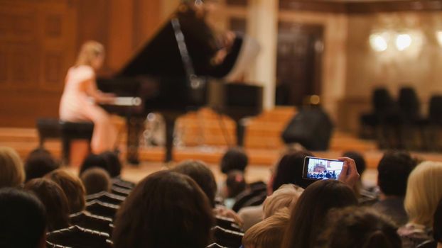 Spectators in concert hall during performing piano girl- people shooting performance on smartphone