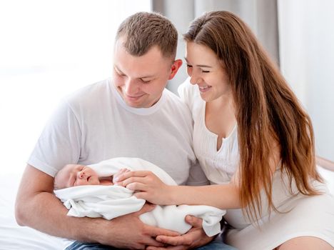 Beautiful young family with newborn daughter in the bed with daylight. Adorable infant baby girl sleeping in hands of her mother and father at home and parents looking at her with smile