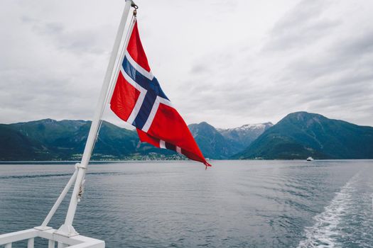 Norwegian flag hanging on the railing of the ship and waving above the water. Norvegian fjord with a flag. Ferry trip in Norway. Norway Flag on sea and mountains background