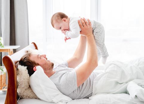 Young father lying in the bed with little son in the sunny morning time and holding kid in his hands. Man with his child baby boy resting in the bedroom. Happy parenthood