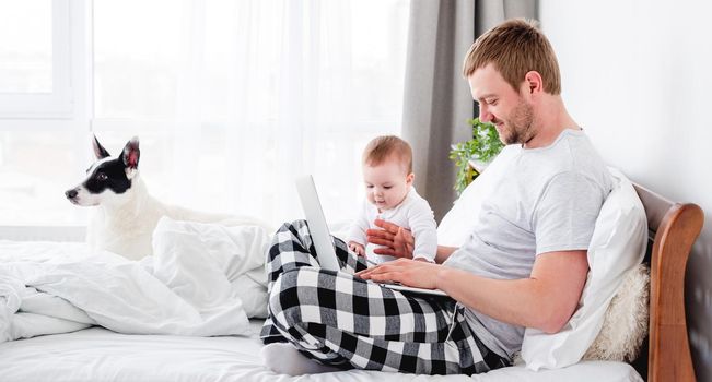 Young father sitting in the bed with little son and cute dog and working on notebook. Man freelancer staying in the bedroom with his child and pet. Guy with kid and doggy in the morning time