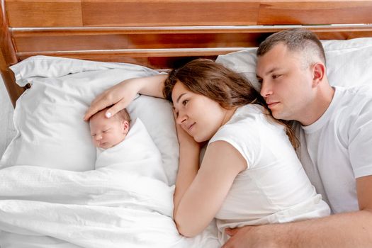 Beautiful young family lying in the bed with newborn daughter together and looking at her. Adorable infant baby girl sleeping close to her mother and father hugging each other