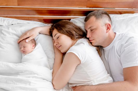 Beautiful young family lying in the bed with newborn daughter and looking at her. Adorable infant baby girl sleeping close to her mother and father