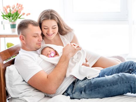 Young parents lying in the bed with newborn daughterand smiling. Adorable infant baby girl sleeping in hands of her mother and father at home