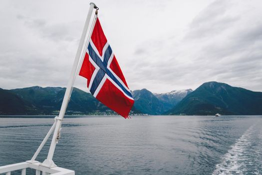 Norwegian flag hanging on the railing of the ship and waving above the water. Norvegian fjord with a flag. Ferry trip in Norway. Norway Flag on sea and mountains background