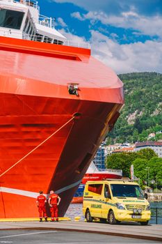 Two Norwegian paramedics in red uniforms are resting near an ambulance parked in a port near a large ship. Theme healthcare and medicine in Norway, Bergen July 28, 2019