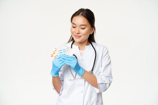 Smiling asian doctor showing medical pills, advertising vitamins, holding drugs with sterile gloves, wearing medical uniform, white background