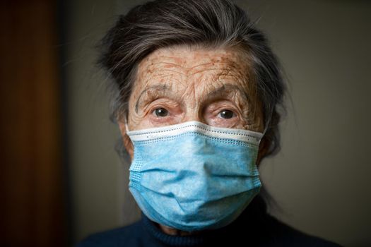 Elderly Caucasian woman with wrinkles on face, gray hair, wearing medical mask, looks attentively at camera, feeling need care and support. Topic safe communication, stay at home during diseases