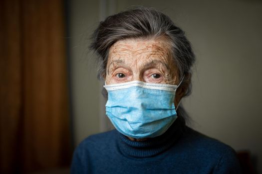 Elderly Caucasian woman with wrinkles on face, gray hair, wearing medical mask, looks attentively at camera, feeling need care and support. Topic safe communication, stay at home during diseases
