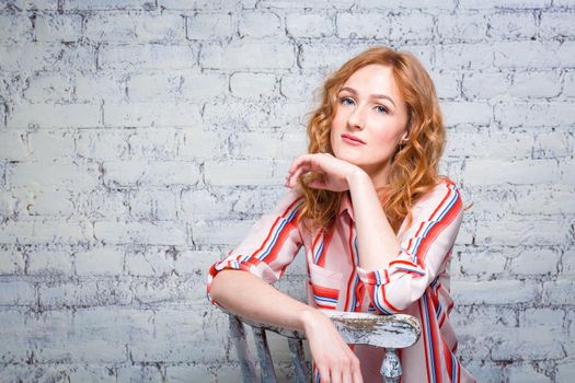 portrait Beautiful young woman student with red curly hair and freckles on her face sitting on a wooden chair on a brick wall background in gray. Dressed in a red striped shirt