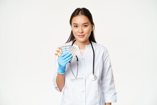 Healthcare and medical concept. Asian female doctor showing pills, vitamins in rubber sterile gloves, standing in uniform over white background