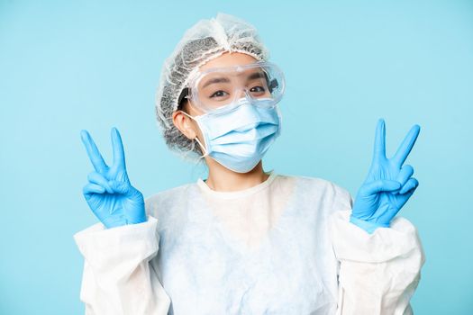 Happy asian female doctor, nurse in personal protective equipment, face mask and sterile gloves, smiling and showing peace, v-signs, blue background