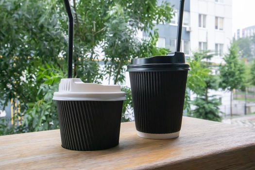 Two black paper coffee cups with lids outdoors in summer in sunny weather on a wooden table of a cafe, coffee shop or restaurant.