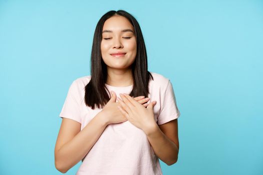 Smiling korean woman dreaming, holding hands on heart and close eyes, cherish memory in her soul, standing over blue background
