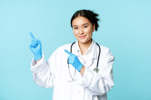 Smiling female doctor, physican in medical uniform and sterile gloves, pointing fingers left at promo, clinic logo, standing over blue background