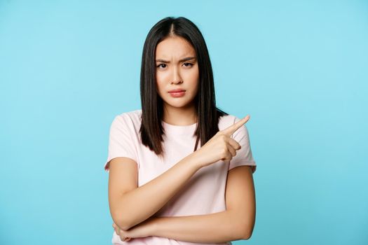 Angry displeased asian woman pointing right, furrow eyebrows in disapproval, standing in t-shirt over blue background
