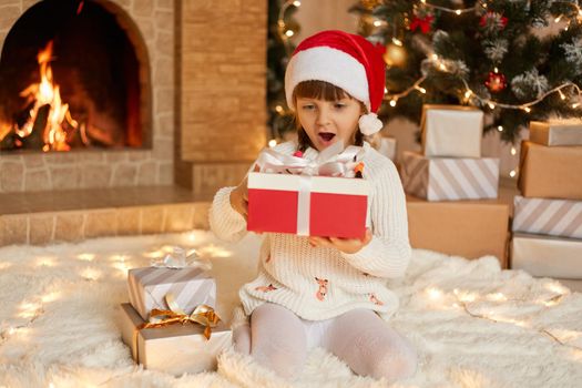 Surprised little girl wearing santa Claus hat and white warm sweater sitting on floor with gift box, look at her present with big eyes and opened mouth, being excited and astonished.
