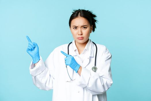 Angry female doctor or nurse, pointing fingers at upper left corner with disapproval, sulking and furrow eyebrows, standing in medical uniform, blue background