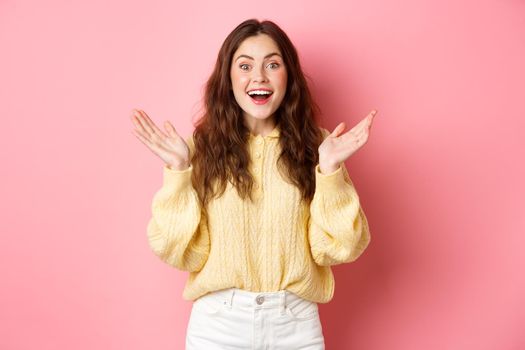 Happy beautiful girl gasping amazed, clap hands and smiling, looking surprised, receive great news, being praised, standing against pink background