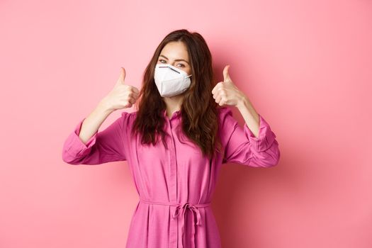 Covid-19, pandemic and lifestyle concept. Cheerful girl shows thumbs up, wears medical respirator as preventive measure from corona, pink background