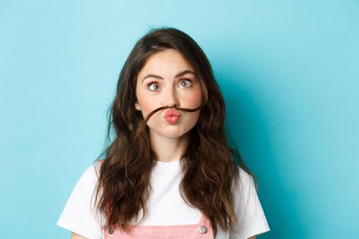 Close up portrait of funny silly girl playing, making squinting face and moustache with hair strand above lips, standing over blue background