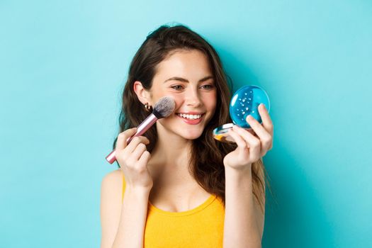 Beauty. Modern girl with candid smile applying make up with brush, looking in pocket mirror, standing over blue background