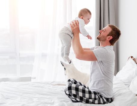 Young father sitting in the bed and holding little son in his hands in the room with sunlight. Man staying in bedroom with his child. Guy with kid in morning time