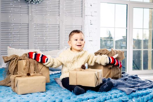little funny playful boy a child sits on a bed on Christmas day with gift boxes in white wool knitted sweater and big bright mittens on it and laughs out loud. In interior there is a festive decor
