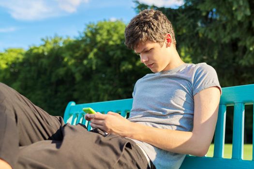 Serious guy teenager using smartphone, having rest, sitting on bench in park